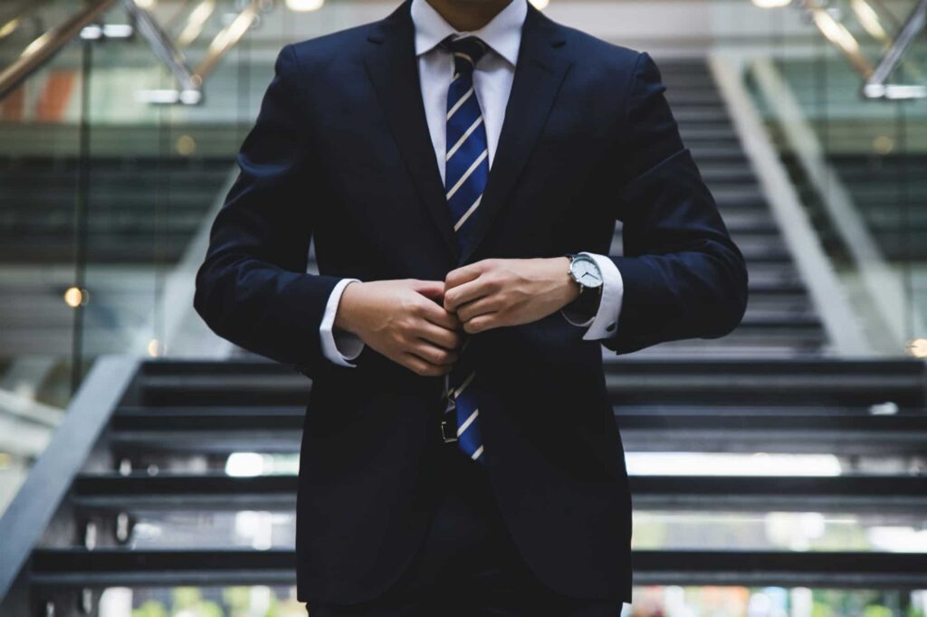 A businessman in a dark suit adjusting his blue striped tie while standing on a staircase in an office
