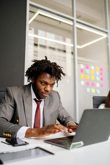 A Black man in a grey suit and red tie types on a laptop at an office desk with a glass partition behind him