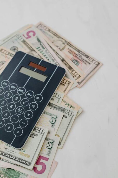 A dark blue calculator resting on top of a scattered pile of US dollar bills on a light surface