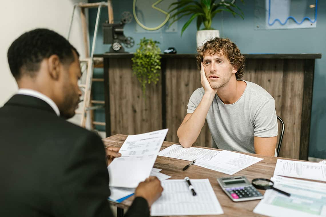 A man in a suit reviews paperwork at a desk across from a stressed consumer looking overwhelmed by debt