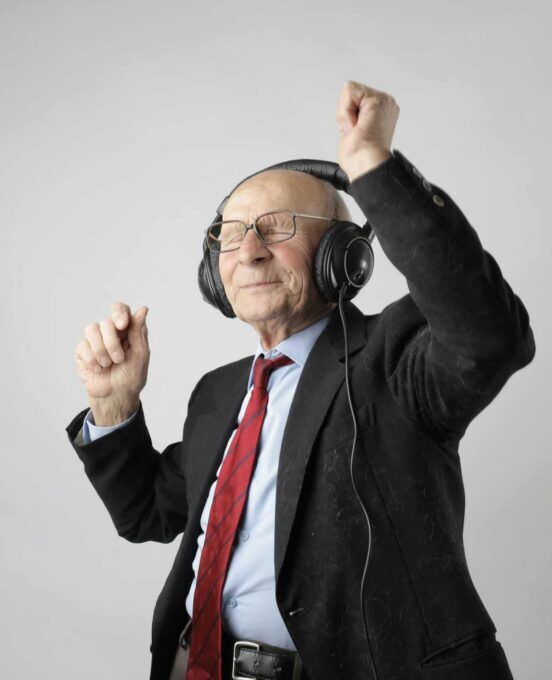 An older man wearing a suit and red tie dances happily with large black headphones against a gray background