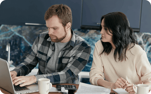 A man typing on a laptop and a woman looking on, sitting at a table with papers, a calculator, and cups